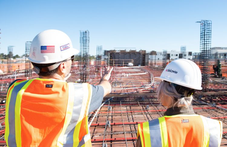 two people talking and standing outside at a construction job site wearing safety vests and hard hats